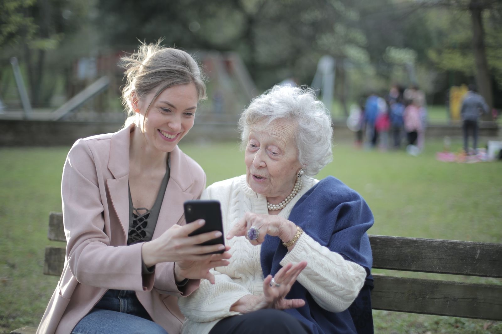 Filha mostrando algo no celular para a mãe idosa, sentadas em um banco de praça